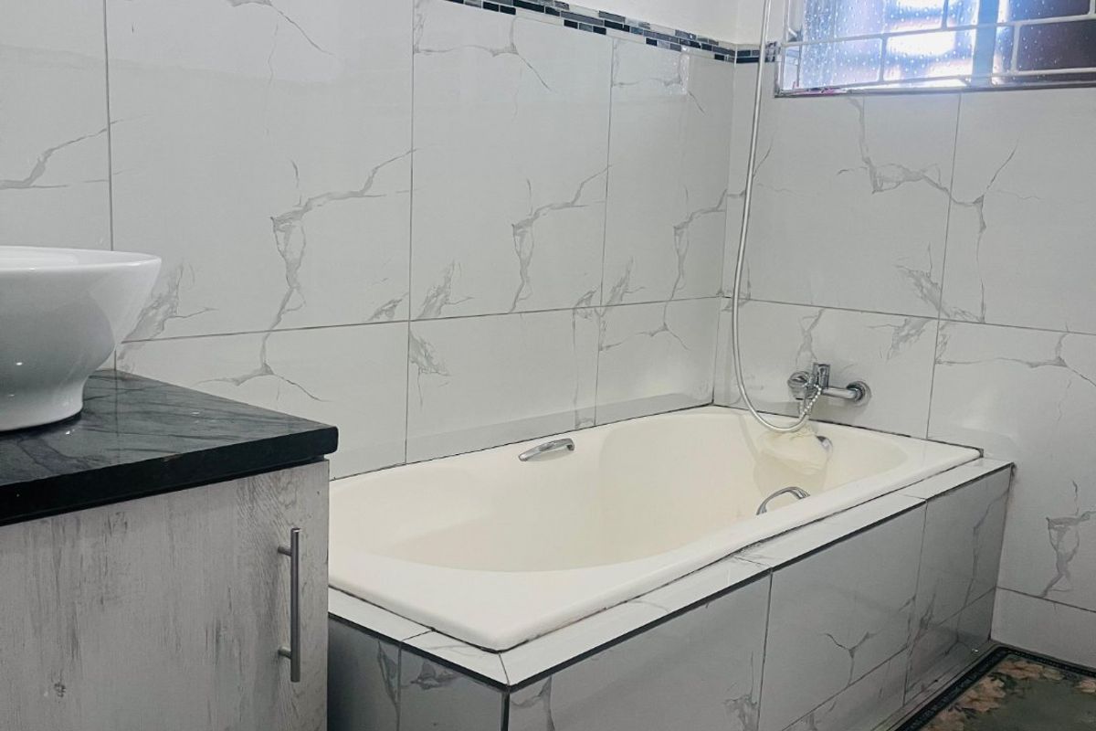 A modern bathroom featuring a built-in bathtub, marble-look wall tiling, a vanity cabinet with a vessel sink, and a window with security bars.