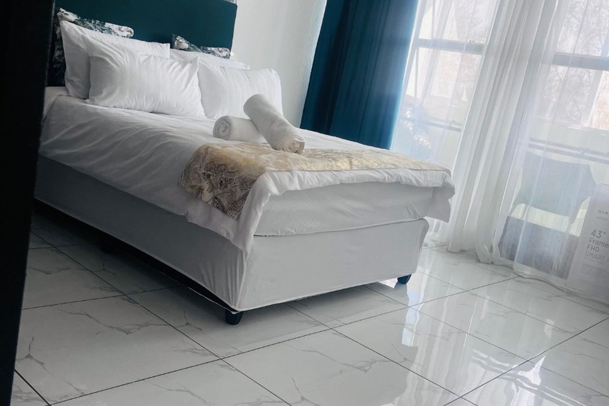 A bright bedroom featuring polished marble-look floor tiles, large windows with curtains, and access to an outdoor balcony area.