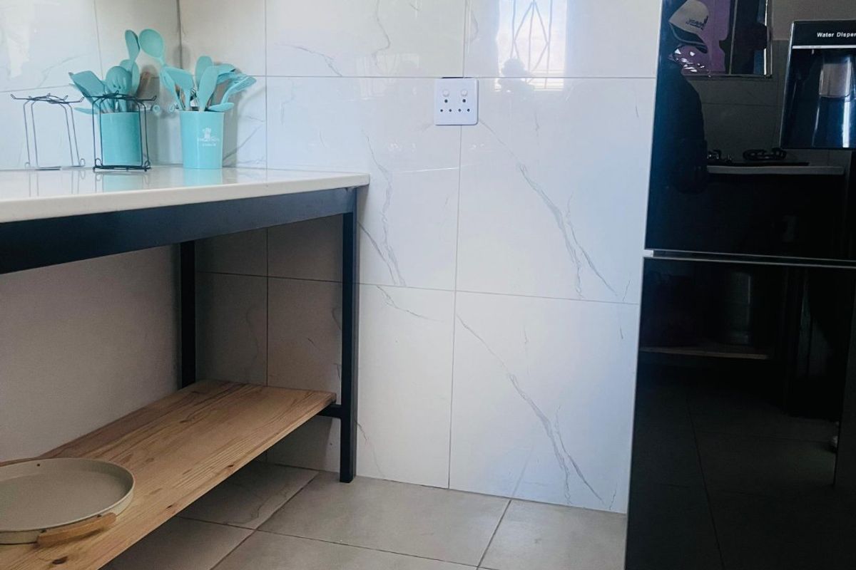 A modern kitchen area featuring marble-look wall tiling, open wooden shelving, and a sleek metal-framed workbench with tiled flooring.