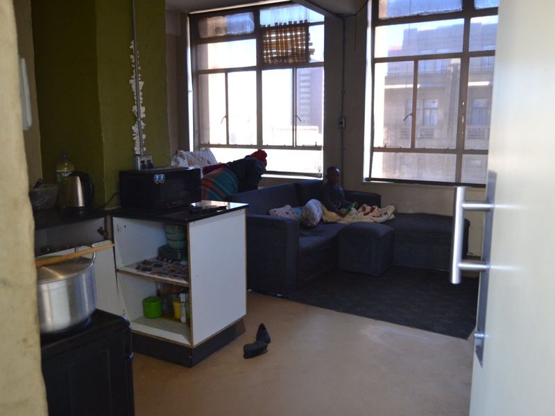 Open-plan living area featuring large windows, built-in kitchen cabinetry, and a combined lounge space in need of cosmetic updates.