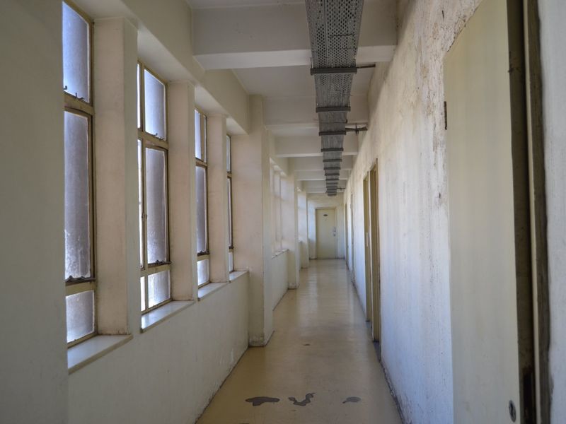 A long, vacant apartment hallway featuring exposed cable trays, multiple windows, and worn wall and floor finishes requiring maintenance.