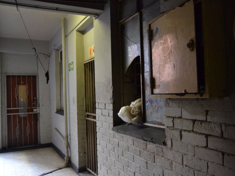 A dimly lit apartment hallway featuring exposed brick walls, tiled flooring, and security gates, showing signs of significant wear and maintenance requirements.