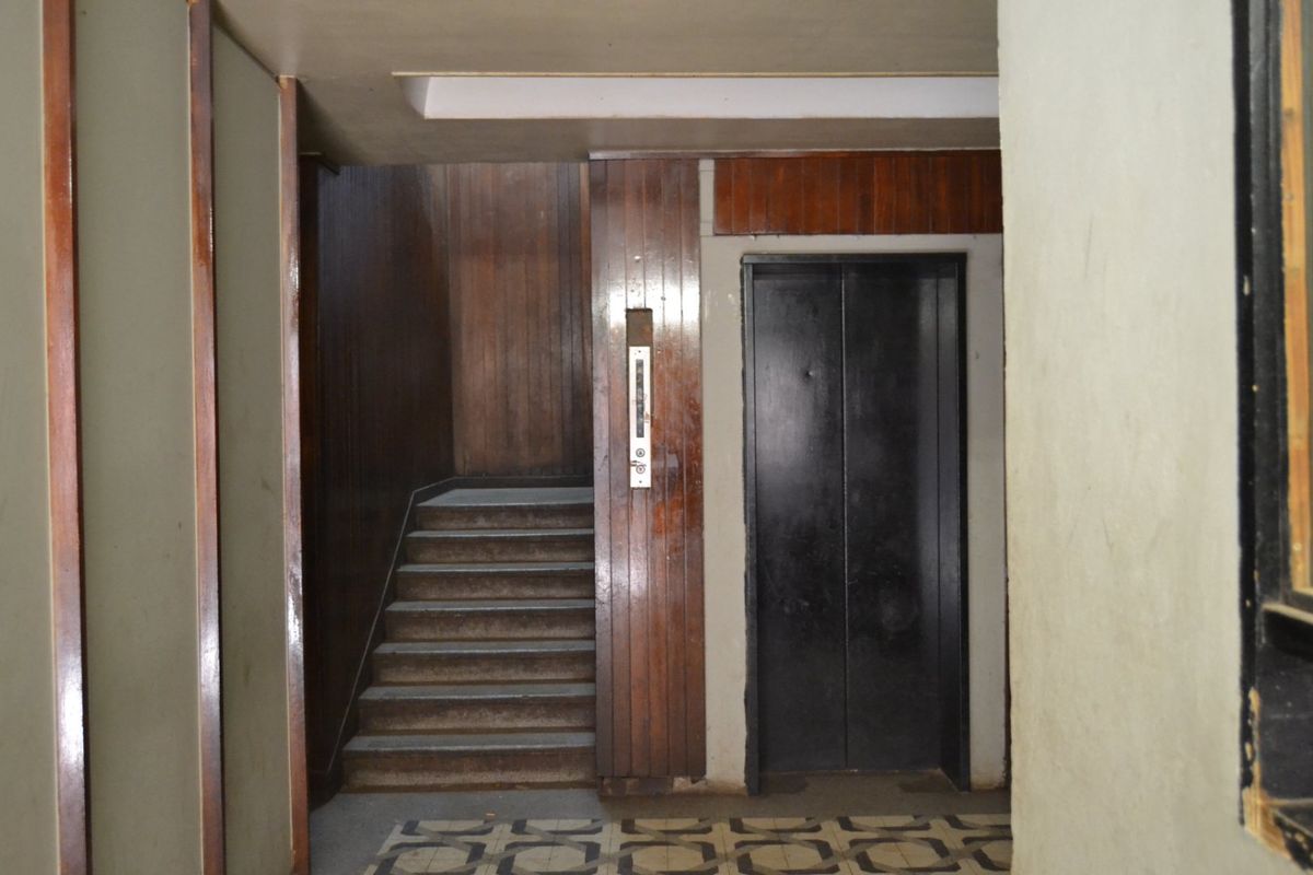 A dated apartment building hallway featuring a functional elevator, a staircase with wood paneling, and original patterned floor tiles.