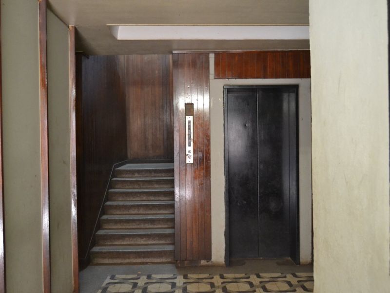 A dated apartment building hallway featuring a functional elevator, a staircase with wood paneling, and original patterned floor tiles.
