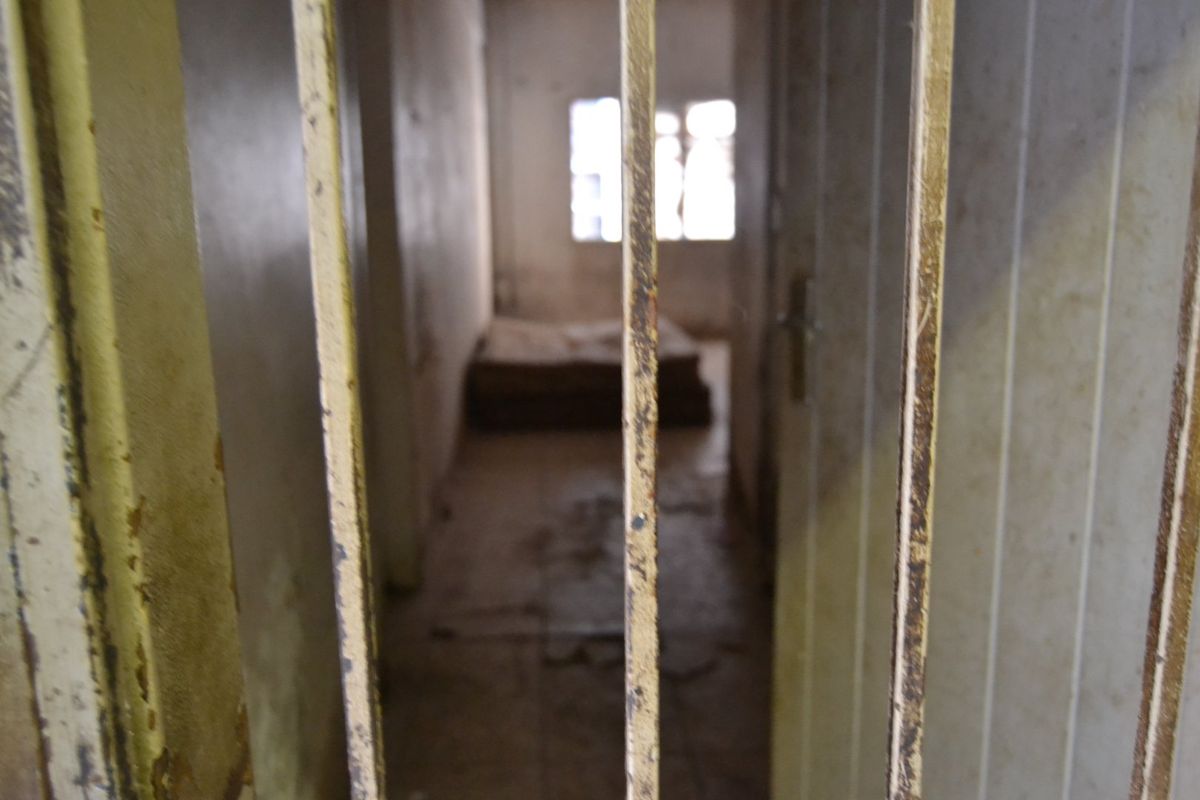 A dilapidated bedroom space viewed through a rusted metal security gate, showing significant wear to walls and flooring.