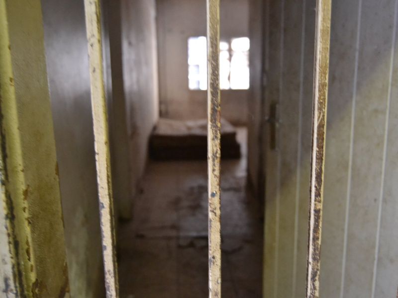 A dilapidated bedroom space viewed through a rusted metal security gate, showing significant wear to walls and flooring.