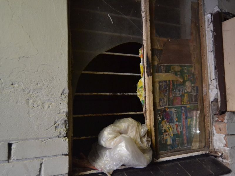 A close-up view of a severely damaged window frame with broken glass and exposed security bars, showing significant deterioration of the surrounding wall structure.