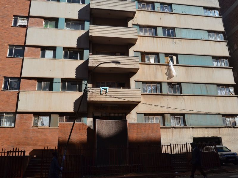 Front elevation of a multi-story brick apartment building featuring balconies and metal security fencing at the street level.
