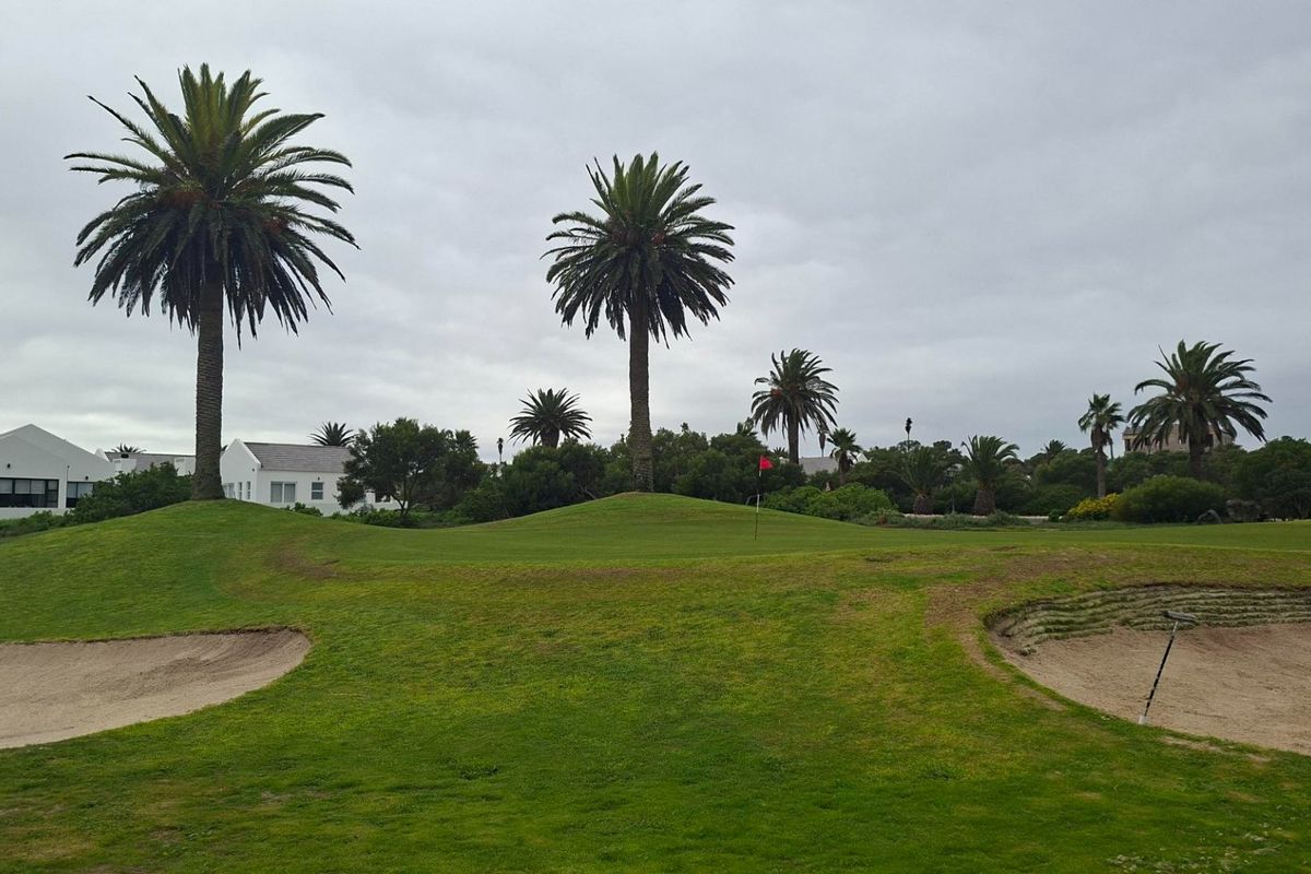 A scenic view of a well-maintained golf course featuring sand bunkers, manicured greens, and mature palm trees under an overcast sky.