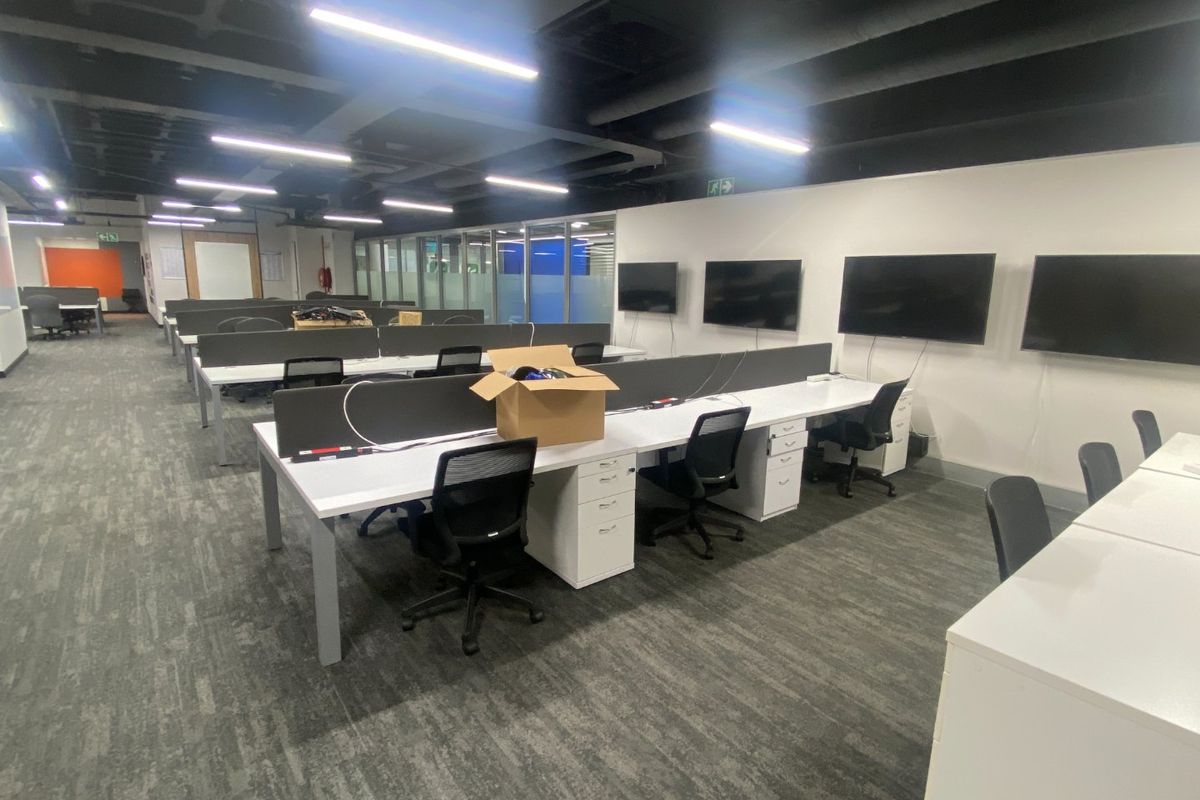 A modern open-plan commercial office space featuring exposed ceilings, linear lighting, carpeted flooring, and integrated wall-mounted screens for collaborative work.