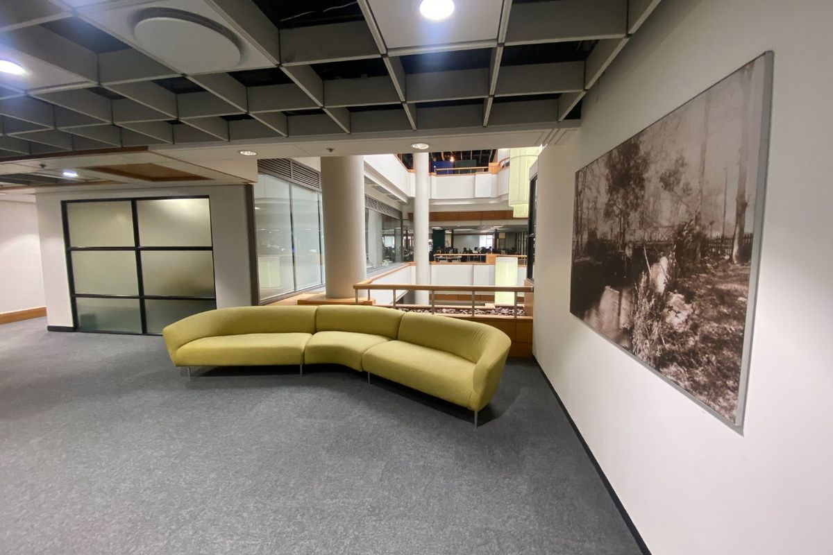 Commercial office lobby or breakout area featuring a suspended ceiling grid, glass partitions, and an open atrium view with structural columns.