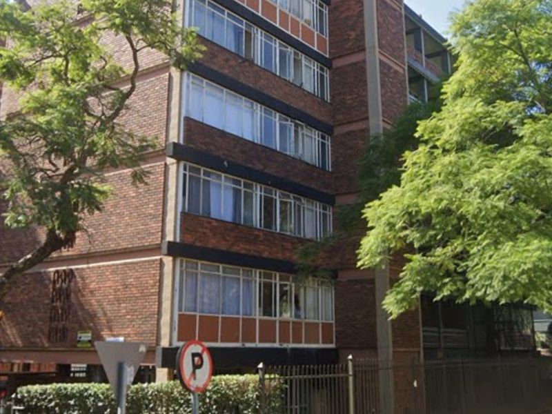 A multi-story brick apartment building featuring metal-framed windows and perimeter fencing, viewed from the street level.