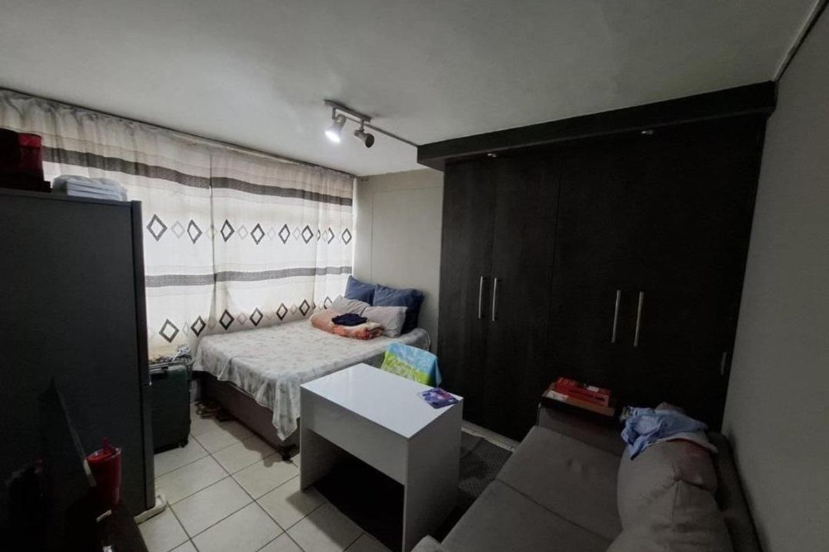 A compact bedroom featuring built-in dark wood cupboards, tiled flooring, and a window with curtains, offering functional storage and living space.
