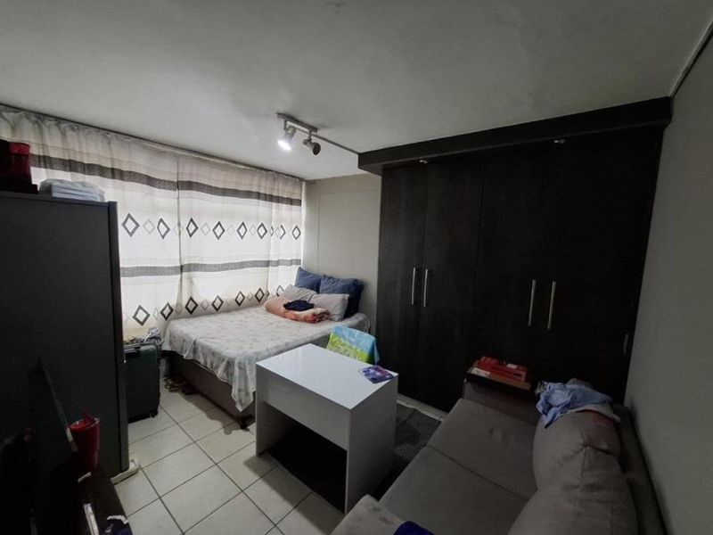 A compact bedroom featuring built-in dark wood cupboards, tiled flooring, and a window with curtains, offering functional storage and living space.