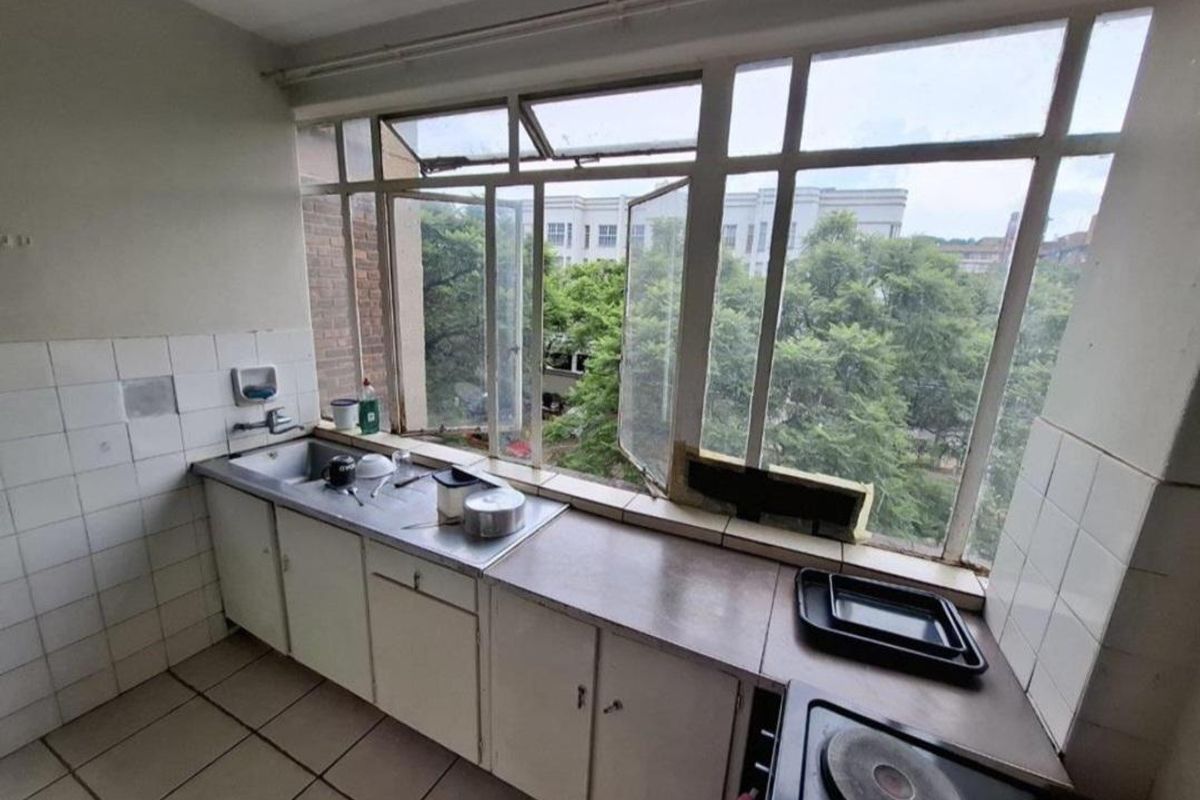 A dated kitchen featuring built-in cabinetry, a stainless steel sink, and an electric hob, positioned against a large window with views of greenery.
