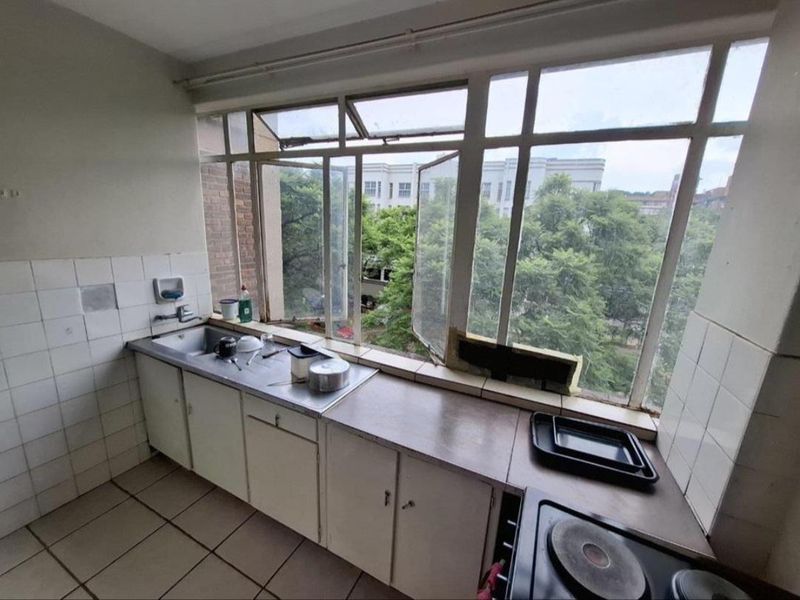 A dated kitchen featuring built-in cabinetry, a stainless steel sink, and an electric hob, positioned against a large window with views of greenery.