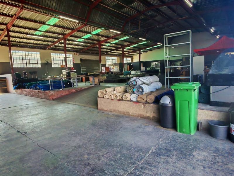 Spacious industrial warehouse area featuring exposed steel roof trusses, translucent skylights, concrete flooring, and a raised brick platform for storage or operational use.