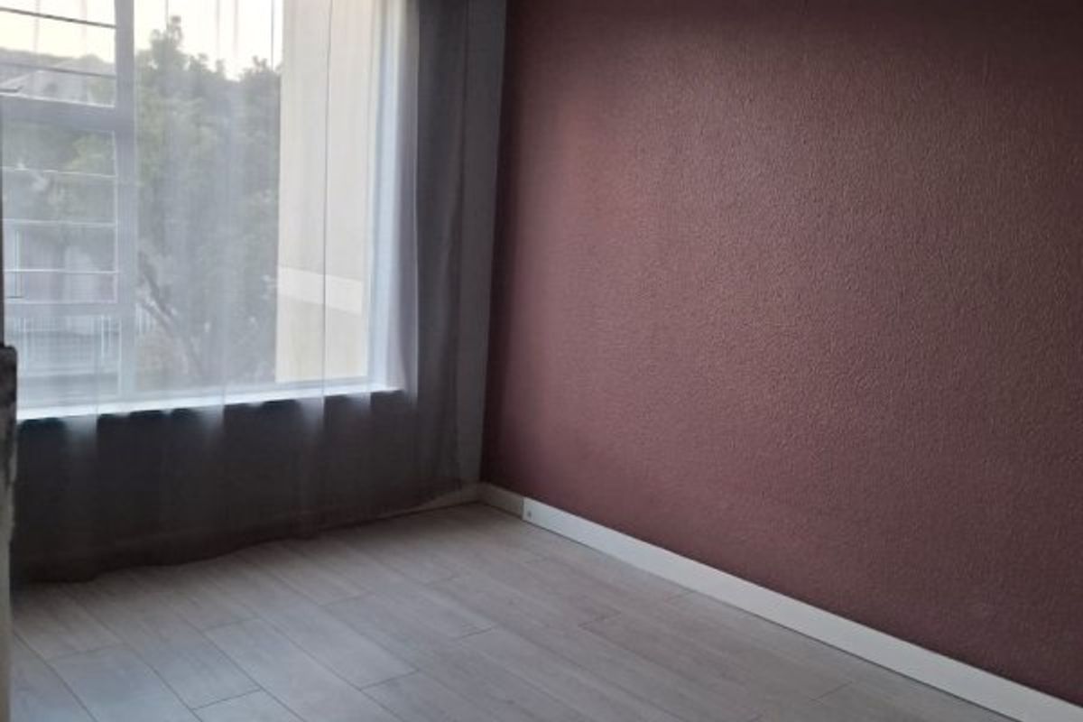 A bright, empty bedroom featuring light-toned laminate flooring, a dark accent wall, and a window fitted with security burglar bars.