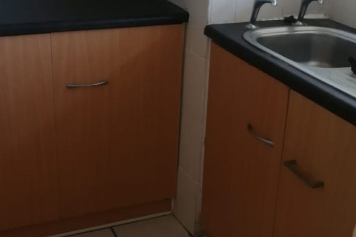 A compact kitchen area featuring wood-look laminate cabinetry, dark countertops, a stainless steel sink with dual taps, and neutral ceramic floor and wall tiling.