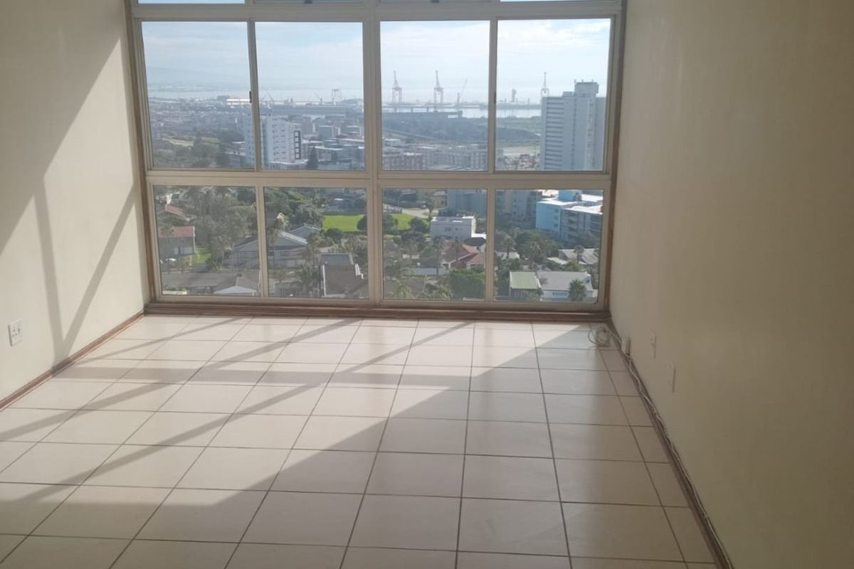An empty, sunlit living area featuring large windows with expansive city and harbour views, finished with neutral ceramic floor tiles.