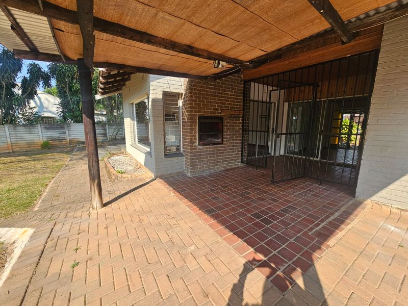 Covered outdoor patio area featuring a built-in braai, brick flooring, and direct access to the garden through security gates.