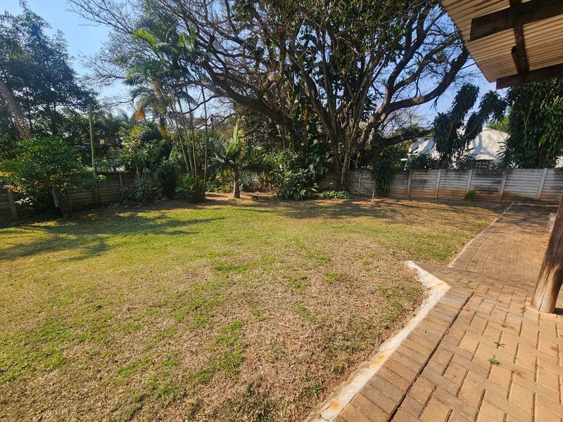 A spacious rear garden featuring a paved patio area, established mature trees, and a secure precast concrete boundary wall.
