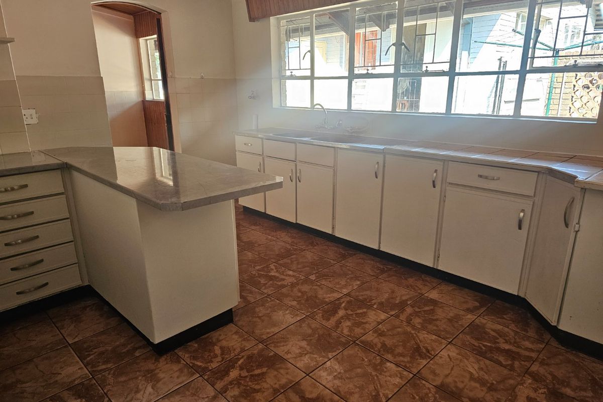 A dated kitchen featuring white built-in cabinetry, laminate countertops, a double basin sink, and tiled flooring with large windows providing natural light.