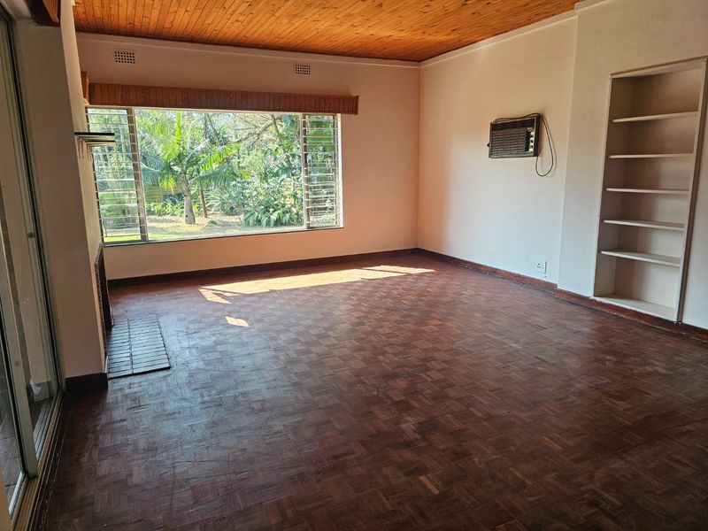 Spacious living room featuring classic parquet flooring, a timber-paneled ceiling, built-in shelving, and a large window overlooking the garden.
