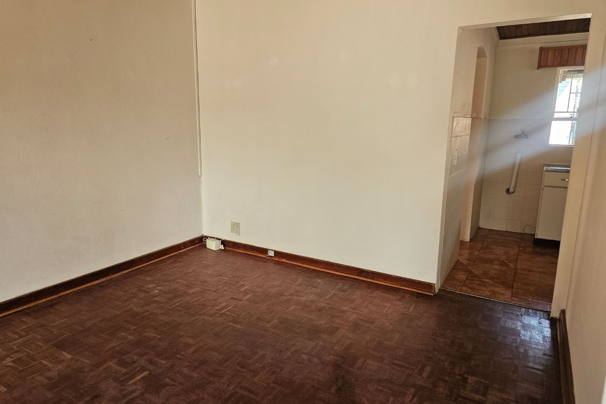 An empty living room featuring original parquet flooring and timber skirting, with an adjacent view into a tiled utility or kitchen area.