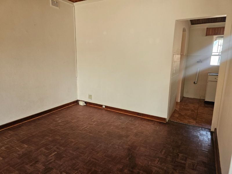 An empty living room featuring original parquet flooring and timber skirting, with an adjacent view into a tiled utility or kitchen area.