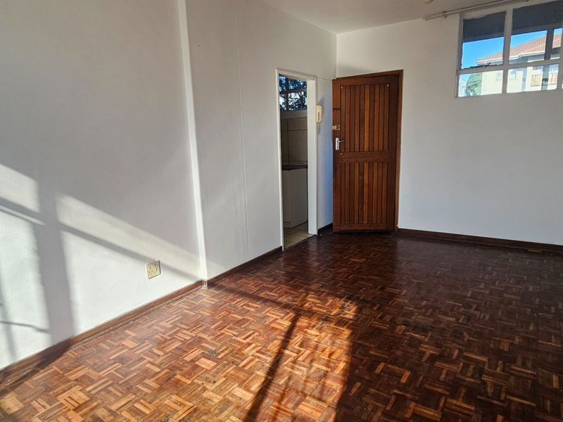 A bright, empty living room featuring classic parquet flooring, a solid timber door, and natural light streaming through a multi-pane window.