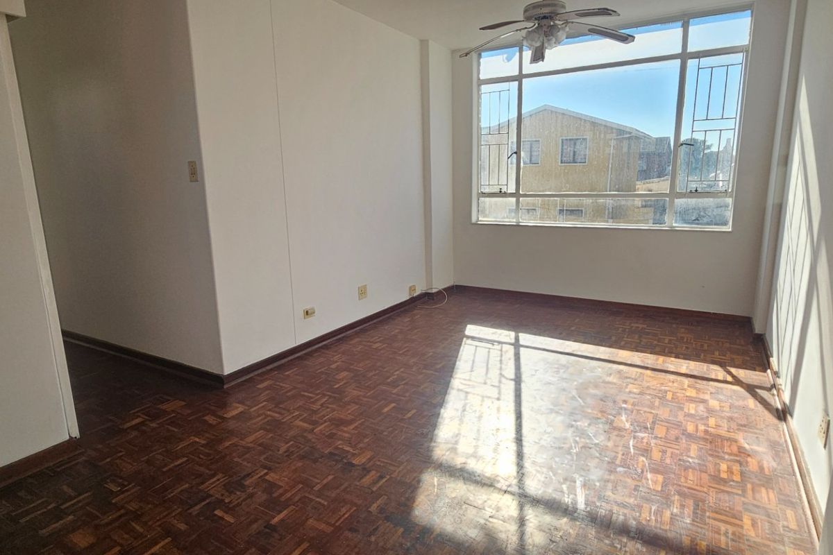 An empty living room featuring original parquet flooring, a ceiling fan, and a large window with security bars, offering natural light.