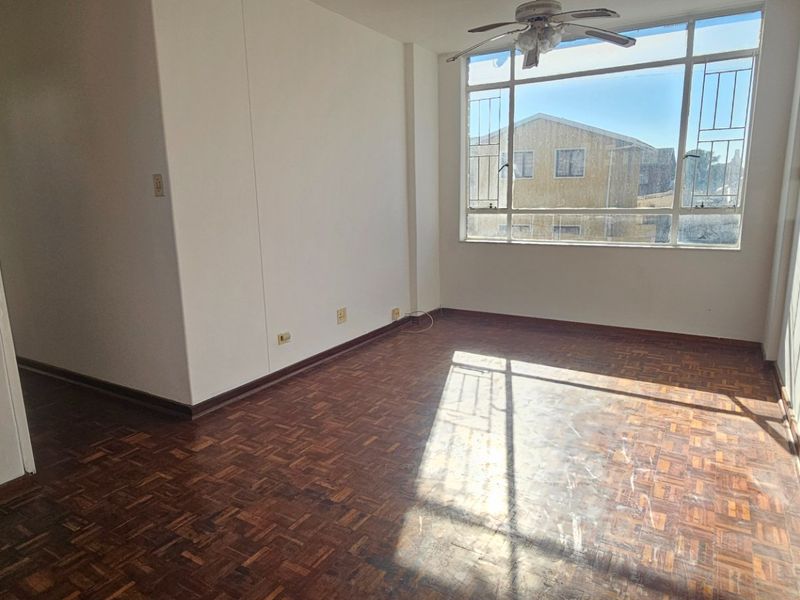 An empty living room featuring original parquet flooring, a ceiling fan, and a large window with security bars, offering natural light.