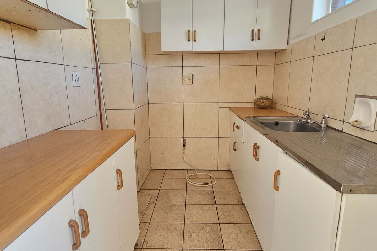 A compact, empty kitchen featuring built-in white cabinetry, laminate countertops, a stainless steel sink, and tiled finishes throughout.