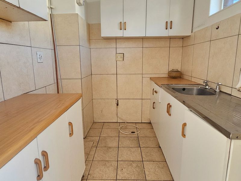 A compact, empty kitchen featuring built-in white cabinetry, laminate countertops, a stainless steel sink, and tiled finishes throughout.