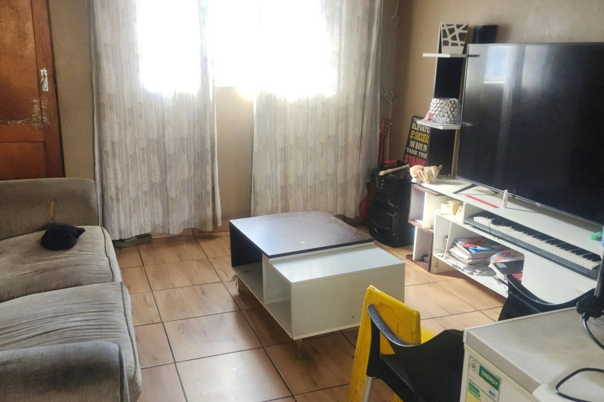 A compact living room featuring tiled flooring and a wooden door, offering a functional space that could benefit from cosmetic updates.