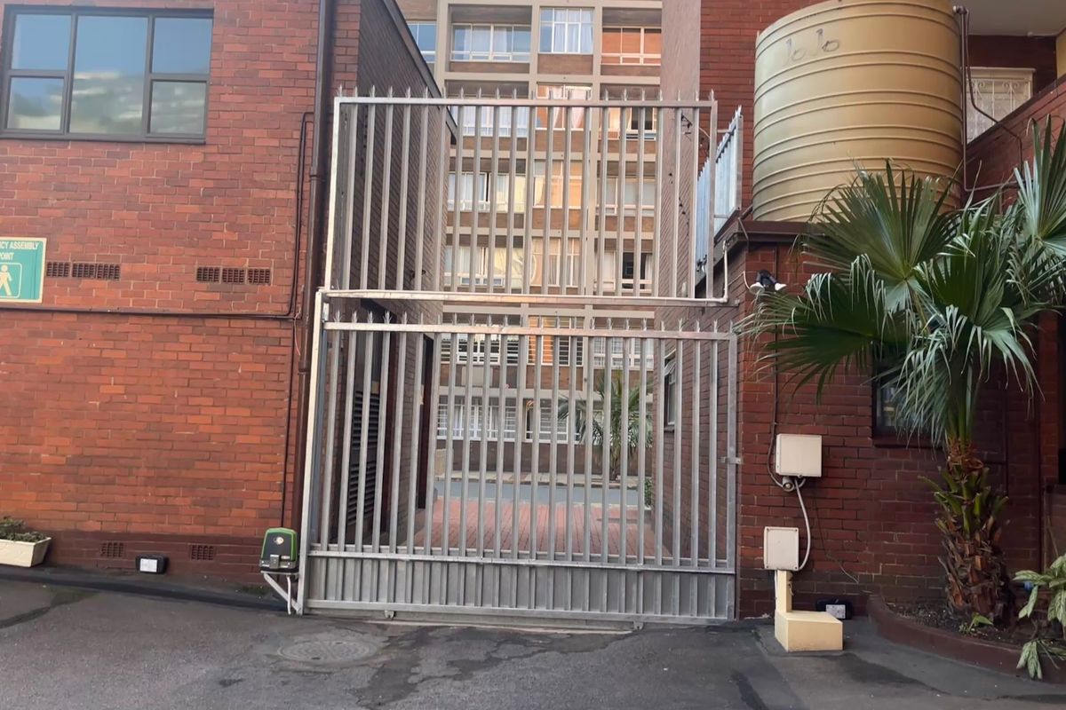 Rear exterior view of the apartment building featuring a secure metal gate, brick facade, and a water storage tank.