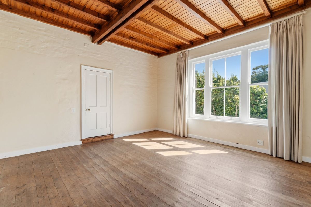 A bright, empty bedroom featuring exposed timber beam ceilings, original timber flooring, and large sash windows overlooking the garden.