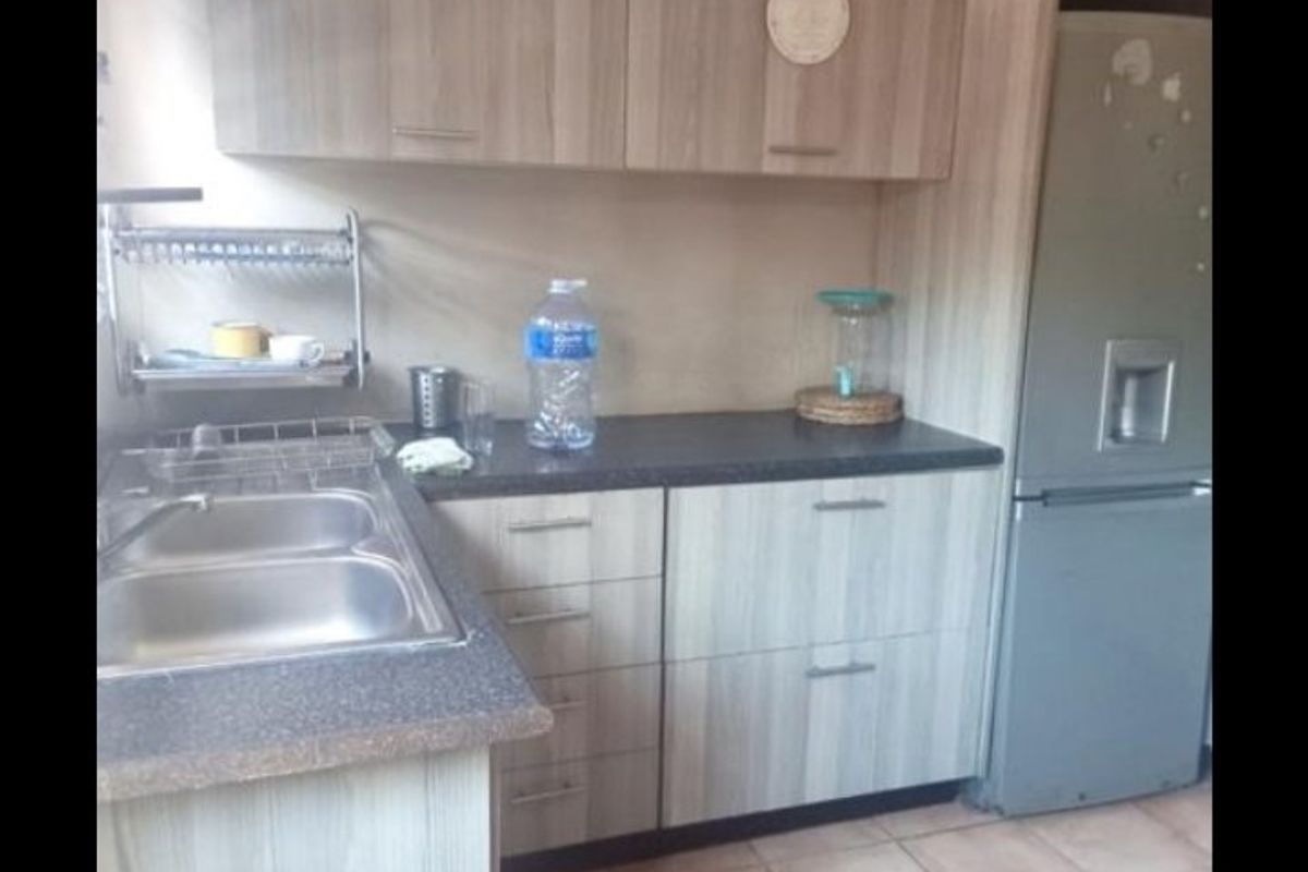A functional kitchen featuring light wood-grain laminate cabinetry, dark countertops, a double bowl stainless steel sink, and ceramic floor tiles.