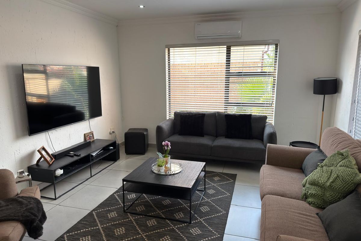 A well-maintained living room featuring tiled flooring, recessed downlighting, and a wall-mounted air conditioning unit with large windows fitted with venetian blinds.