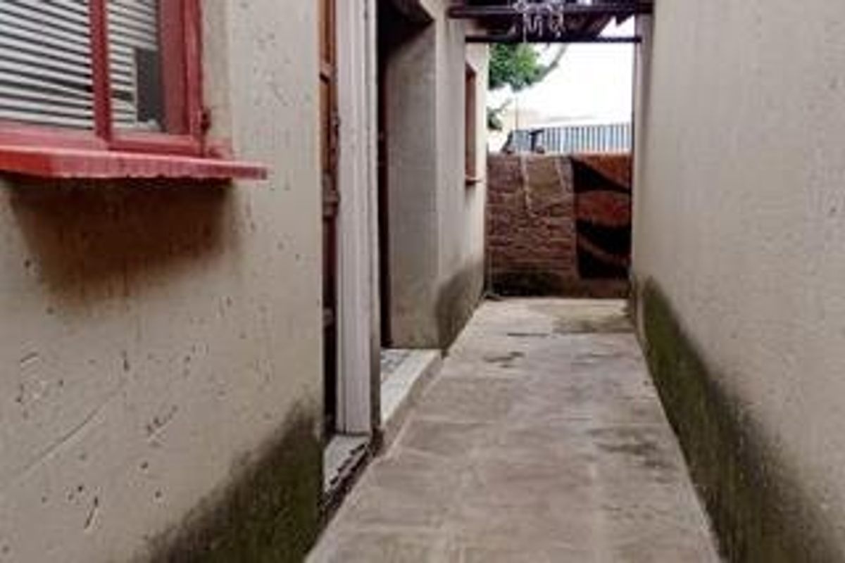A narrow exterior side passage featuring concrete paving and brick boundary wall, showing signs of significant wear and rising damp on the building exterior.