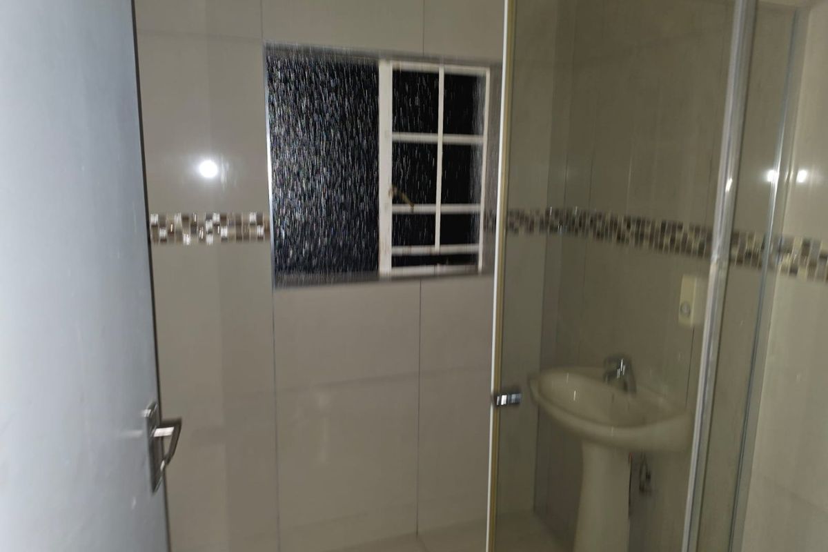 A functional bathroom featuring a pedestal basin, tiled walls with a decorative mosaic border, and a glass-enclosed shower area.