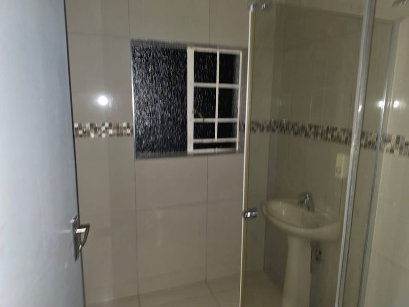 A functional bathroom featuring a pedestal basin, tiled walls with a decorative mosaic border, and a glass-enclosed shower area.