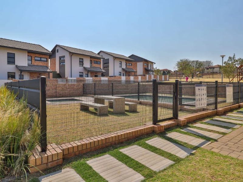 A communal pool area featuring a swimming pool enclosed by mesh fencing, brick boundary walls, and a paved walkway within a townhouse complex.