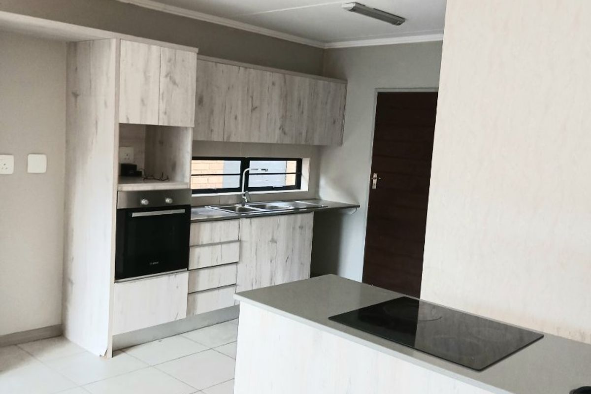 A clean, modern kitchen featuring light wood-grain cabinetry, stone-look countertops, a built-in oven, and a central island with an integrated electric hob.
