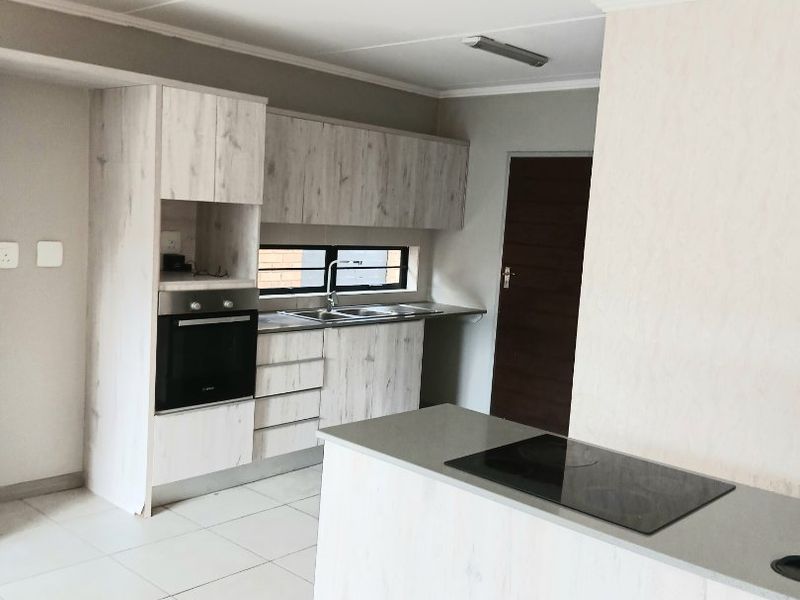 A clean, modern kitchen featuring light wood-grain cabinetry, stone-look countertops, a built-in oven, and a central island with an integrated electric hob.