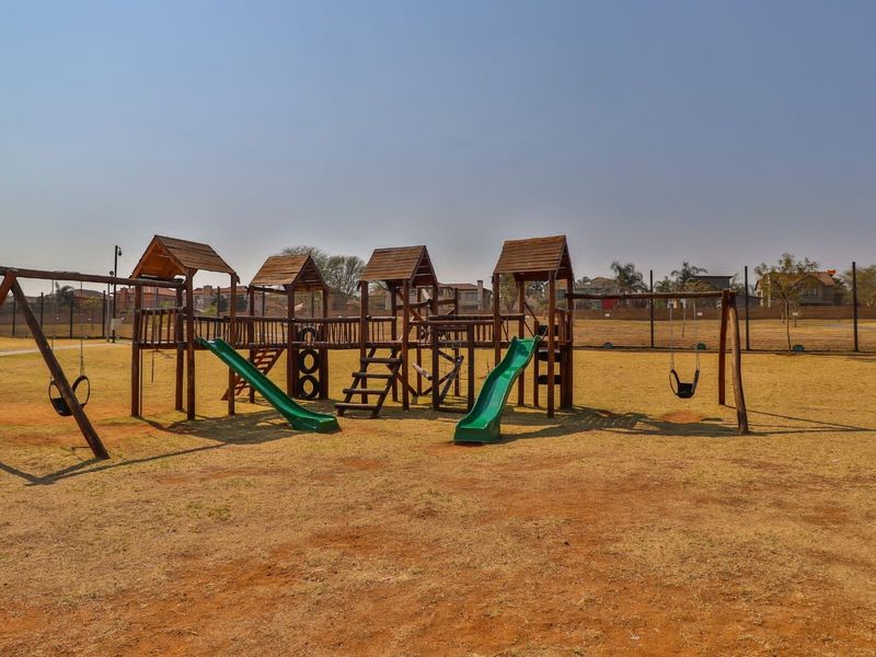A communal outdoor garden area featuring a large wooden playground structure with slides and swings, set within a fenced residential estate.