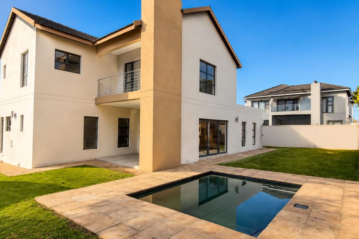 Rear view of a modern two-story house featuring a swimming pool, stone patio area, upper-level balcony, and a well-maintained garden.
