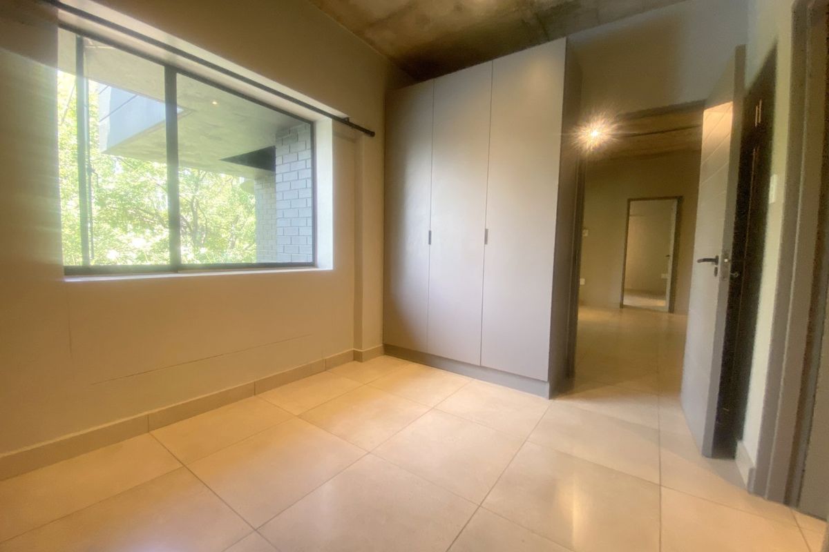 A bright, empty bedroom featuring modern built-in cupboards, large windows, tiled flooring, and an industrial-style exposed concrete ceiling.
