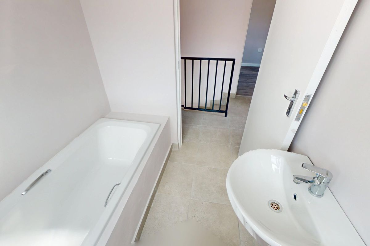 A clean, modern bathroom featuring a built-in bathtub and pedestal washbasin, with neutral tiled flooring and direct access to the staircase landing.